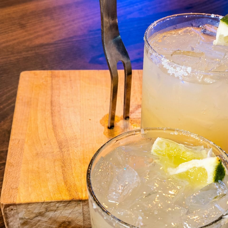Two citrus cocktails with salted rims and lime wedges sit on a wooden serving board under colorful restaurant lighting.
