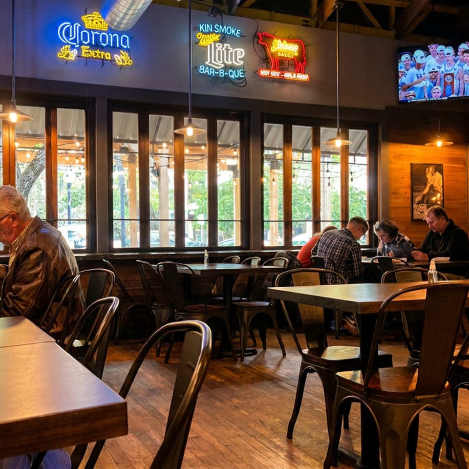 Warmly lit barbecue restaurant dining room with wooden tables, metal chairs, neon beer signs, televisions, and several diners.