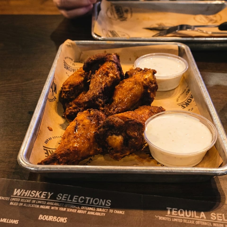 Buffalo chicken wings with two cups of creamy dipping sauce on a metal tray beside a whiskey drink and menu.
