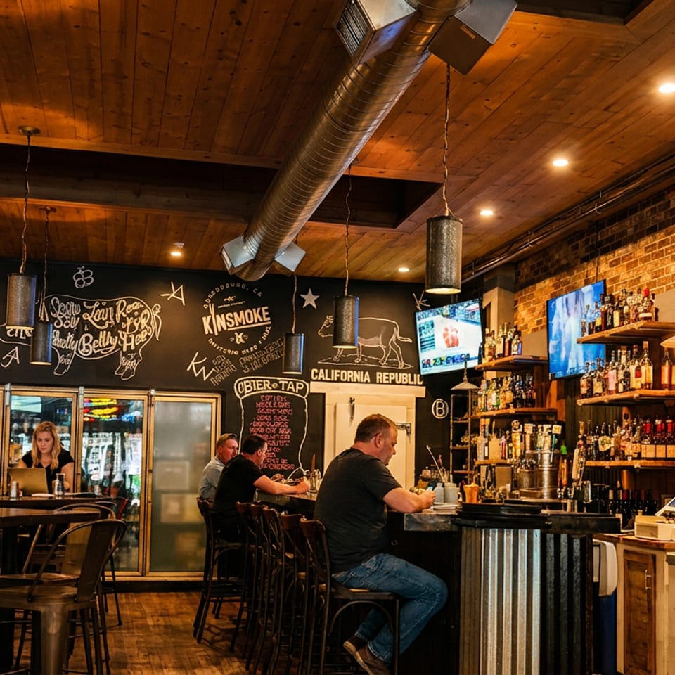 Warmly lit restaurant bar with exposed brick, wood ceilings, chalkboard art, liquor shelves, tables, and patrons seated at the counter.