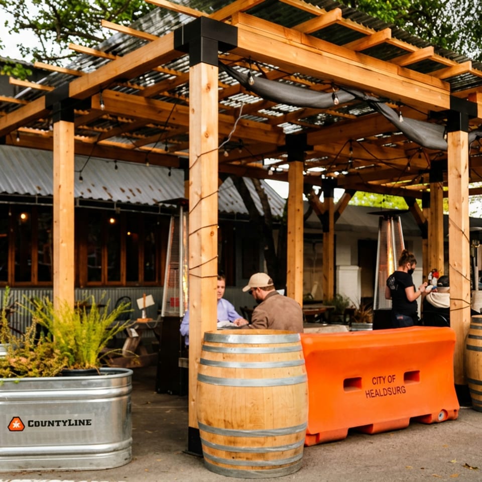 Outdoor restaurant patio beneath a wooden pergola, with diners, wine barrels, metal planters, heaters, and orange City of Healdsburg barriers.