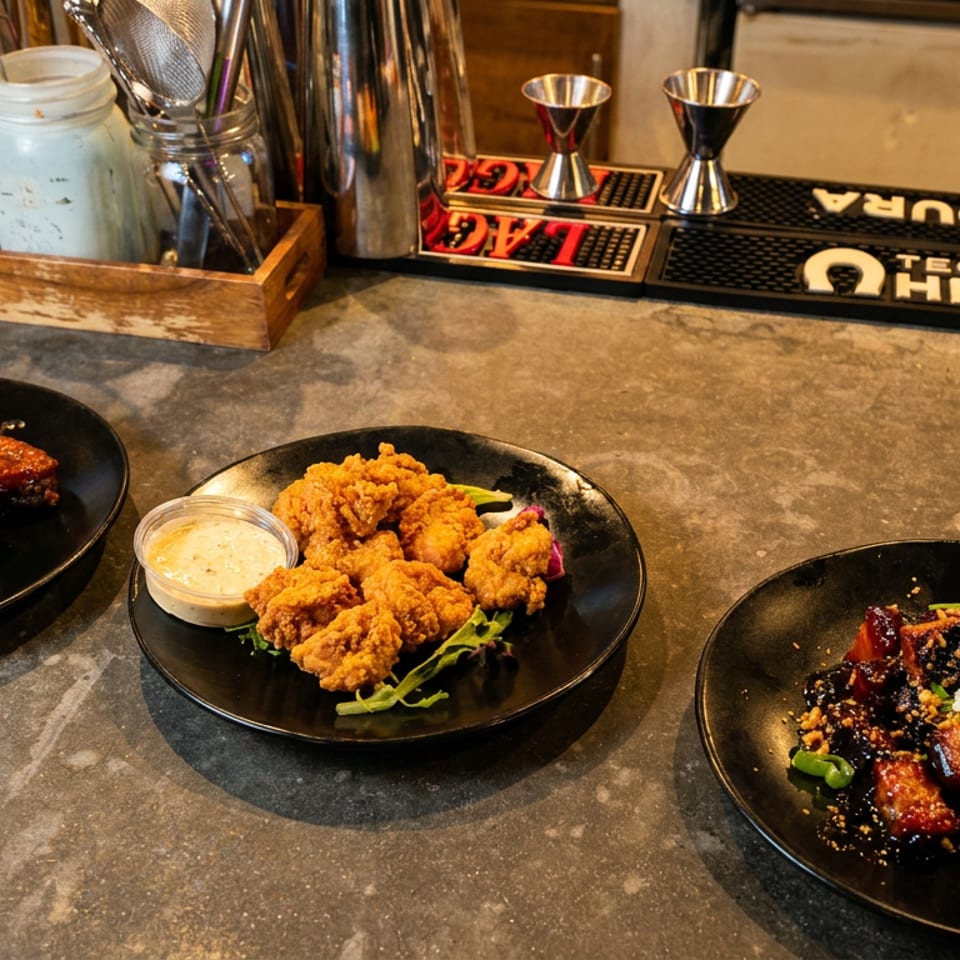 Three black plates hold glazed wings, fried bites with dipping sauce, and saucy garnished pieces beside cocktails on a bar.